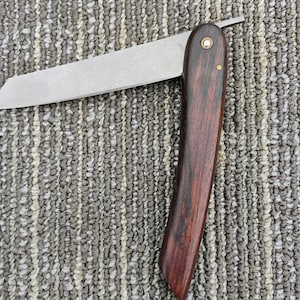 May include: A straight razor with a wooden handle. The handle is dark brown with a reddish hue and has a brass pin at the top. The blade is silver and has a slight curve.