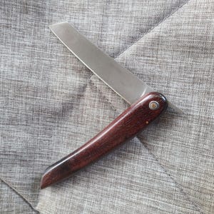 May include: A brown wooden handled pocket knife with a silver blade. The knife is closed and lying on a gray fabric surface.