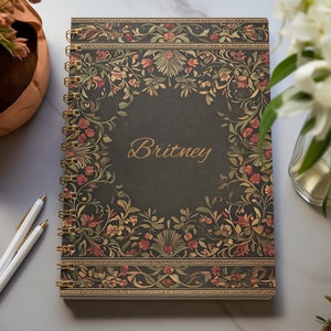 Puede incluir: Una libreta de espiral negra con un diseño floral dorado. El nombre "Britney" está escrito en letras doradas en la portada.