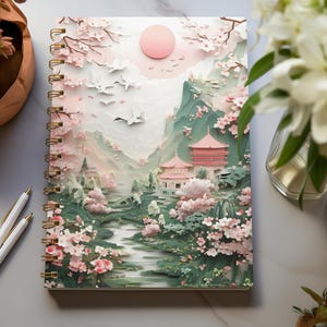 Può includere: Un taccuino a spirale con una copertina in carta rosa e verde che presenta un design di papercraft 3D di un paesaggio giapponese con fiori di ciliegio, montagne, una pagoda e uccelli.