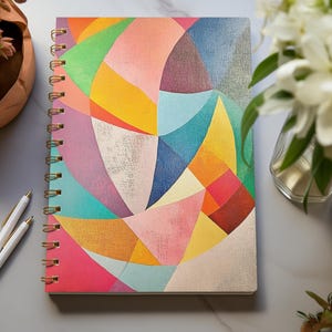 Puede incluir: Un cuaderno de espiral con un diseño geométrico abstracto colorido con tonos de rosa, amarillo, azul, verde y morado. El cuaderno tiene un fondo blanco y una encuadernación en espiral dorada.