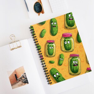 May include: A spiral-bound notebook featuring a repeating pattern of cartoon pickles and pickle jars on a yellow background. The pickles have various facial expressions. The notebook is open on a white surface.