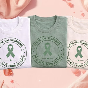 May include: Three t-shirts in white, green, and cream colors, each featuring a circular design with the text "ALPHA GAL SYNDROME" and "TICK-BITE FOOD ALLERGY" around a green ribbon. A pink drink with a straw sits in the foreground.