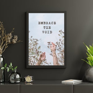 May include: A black framed print with the text "EMBRACE THE VOID" in white. The print features a photo of two hands reaching up towards a blue sky through a field of white flowers.