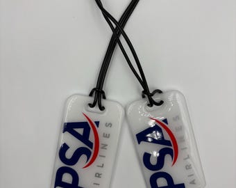 May include: Two white luggage tags with black string loops. Each tag features the PSA Airlines logo in blue, red, and white.