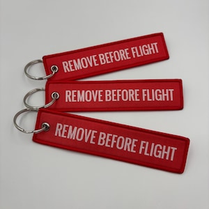 May include: Three red rectangular keychains with the text "REMOVE BEFORE FLIGHT" in white. Each keychain has a silver metal ring attached. The keychains are arranged on a white surface.