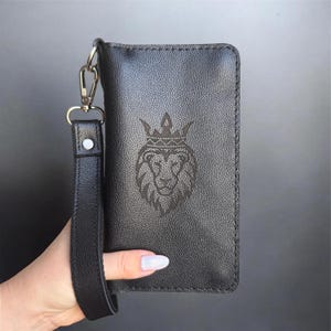 Peut inclure: Portefeuille-bracelet en cuir noir avec fermoir et dragonne argentés. Le portefeuille présente une tête de lion gravée portant une couronne. Le bracelet est tenu par une personne.