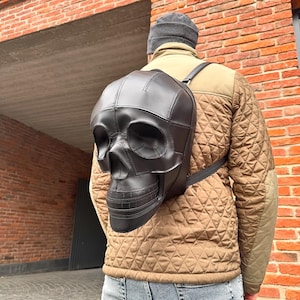 Könnte beinhalten: Ein schwarzer Leder-Schädel-Rucksack mit einem gesteppten Design. Der Rucksack wird von einer Person getragen, die eine braune gesteppte Jacke und eine blaue Jeans trägt.