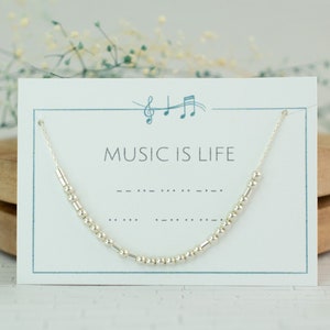 May include: A silver necklace with a Morse code design, displayed on a card that reads "MUSIC IS LIFE." The necklace is delicate, with small beads and bars forming the code. The card has a musical note graphic and is set against a soft, neutral background.