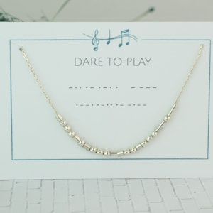 May include: A silver beaded necklace displayed on a white card with the text "DARE TO PLAY" and musical notes. The necklace is strung on a delicate gold chain. The card has a light blue border.