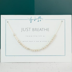 May include: A silver necklace with a Morse code design is displayed on a white card. The card has the words "JUST BREATHE" printed on it. The necklace is made of small, silver beads and is strung on a delicate chain. The background is a teal-colored box.