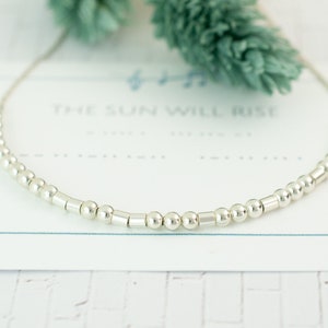 Puede incluir: Un delicado collar de plata con una serie de pequeñas cuentas redondas y barras cilíndricas. El collar se muestra sobre una tarjeta blanca con el texto "THE SUN WILL RISE" en azul claro. Un ramito verde en el fondo.