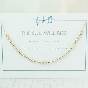 Puede incluir: Un collar de cuentas plateadas con el mensaje en código Morse "THE SUN WILL RISE" mostrado en una tarjeta blanca con un borde azul. El collar está ensartado en una delicada cadena.