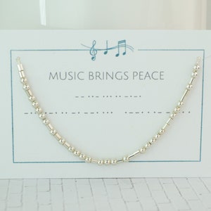 May include: A silver-colored necklace with a Morse code design, displayed on a white card. The card has the text "MUSIC BRINGS PEACE" and musical notes. The necklace is made of small, round beads and longer, cylindrical beads.