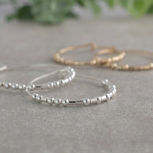 May include: Close-up of several delicate bracelets. Two silver bracelets with small, round beads and a cylindrical accent are in the foreground. Two gold bracelets with similar bead designs are slightly out of focus in the background. The bracelets are on a gray surface.