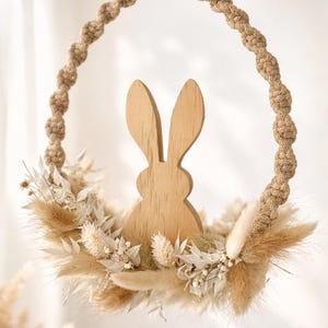 Peut inclure: Couronne de Pâques décorative avec une silhouette de lapin en bois au centre. Le lapin est entouré de fleurs et d'herbes séchées beiges et crème, le tout dans un anneau en macramé. Un élément de décoration intérieure naturel et rustique.