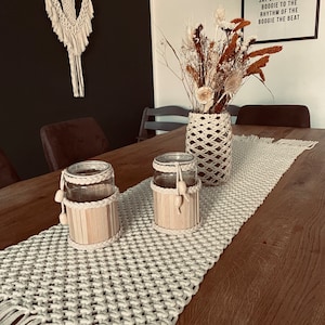 Op de afbeelding: Een witte macramé tafelrunner met franjes aan de randen ligt op een houten tafel met twee glazen potten met houten stokjes erin. Een vaas met gedroogde bloemen staat op de tafel. De muur achter de tafel heeft een zwart-witte poster met de tekst "Hippie to the hip hip-hop and you don't stop the rock it to the bang bang boogie say up jump the boogie to the rhythm of the boogie the beat."