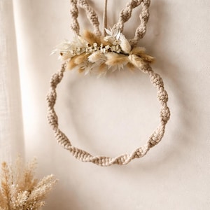 Könnte beinhalten: Ein beigefarbener Makramee-Hasen-Wreath mit Trockenblumen. Der Kranz wird von einer passenden Schnur gehalten. Die Hasenohren sind geknotet, und der Kranz ist mit einem Cluster aus Trockenblumen in Beige- und Weißtönen verziert. Ein dekorativer Artikel.