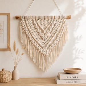 Könnte beinhalten: Cremefarbener Makramee-Wandbehang mit geometrischem Muster, aufgehängt an einem Holzdübel. Die handgefertigte Textilkunst zeichnet sich durch komplizierte Knoten und Fransen aus und verleiht der Inneneinrichtung einen Hauch von Bohème. Ein stilvolles und einzigartiges Stück für jeden Raum.