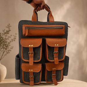May include: A charcoal gray backpack featuring tan leather accents, including a top handle, a horizontal zippered pocket, and four lower pockets with buckled straps. The bag has a structured rectangular shape with brass-toned metal hardware.