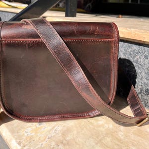 Peut inclure: Un sac &agrave; main en cuir marron avec une longue bandouli&egrave;re r&eacute;glable. Le sac a une fermeture &agrave; rabat et une boucle.