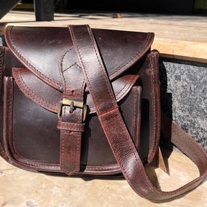 Peut inclure: Un sac &agrave; bandouli&egrave;re en cuir marron avec une fermeture &agrave; rabat et une boucle. Le sac a une seule bandouli&egrave;re et une petite poche sur le devant.