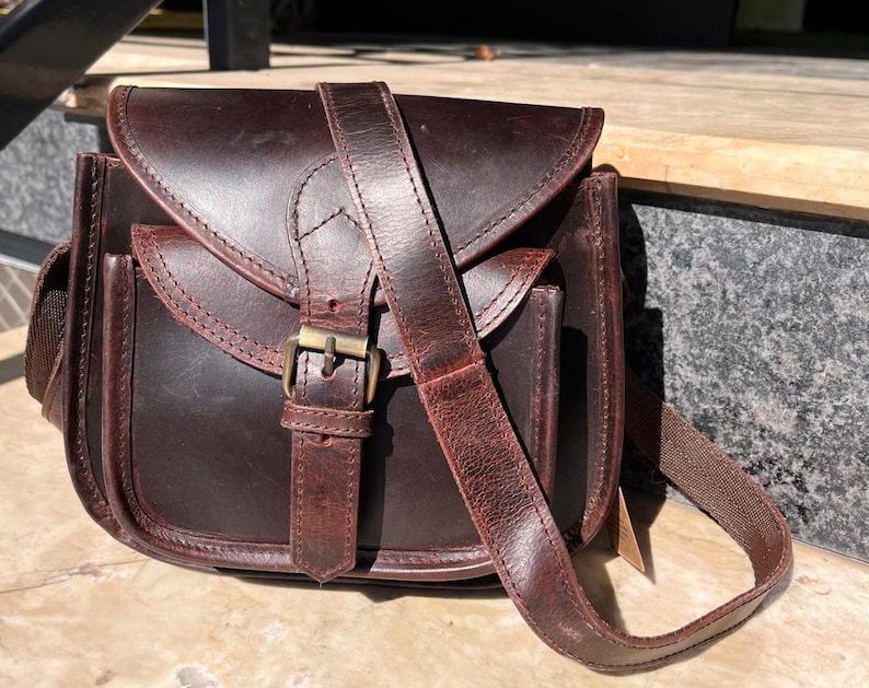 Peut inclure: Un sac &agrave; bandouli&egrave;re en cuir marron avec une fermeture &agrave; rabat et une boucle. Le sac a une seule bandouli&egrave;re et une petite poche sur le devant.