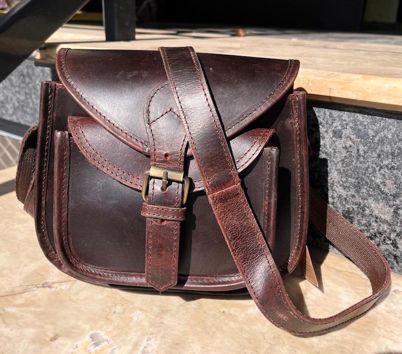 Peut inclure: Un sac &agrave; bandouli&egrave;re en cuir marron avec une fermeture &agrave; rabat et une boucle. Le sac a une poche avant et une longue bandouli&egrave;re r&eacute;glable.