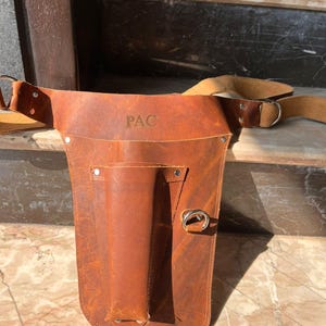 Puede incluir: Un cintur&oacute;n de herramientas de cuero marr&oacute;n con un solo bolsillo y un anillo de metal. El cintur&oacute;n tiene una correa que se coloca sobre el hombro y las palabras "PAC" est&aacute;n grabadas en la parte delantera.