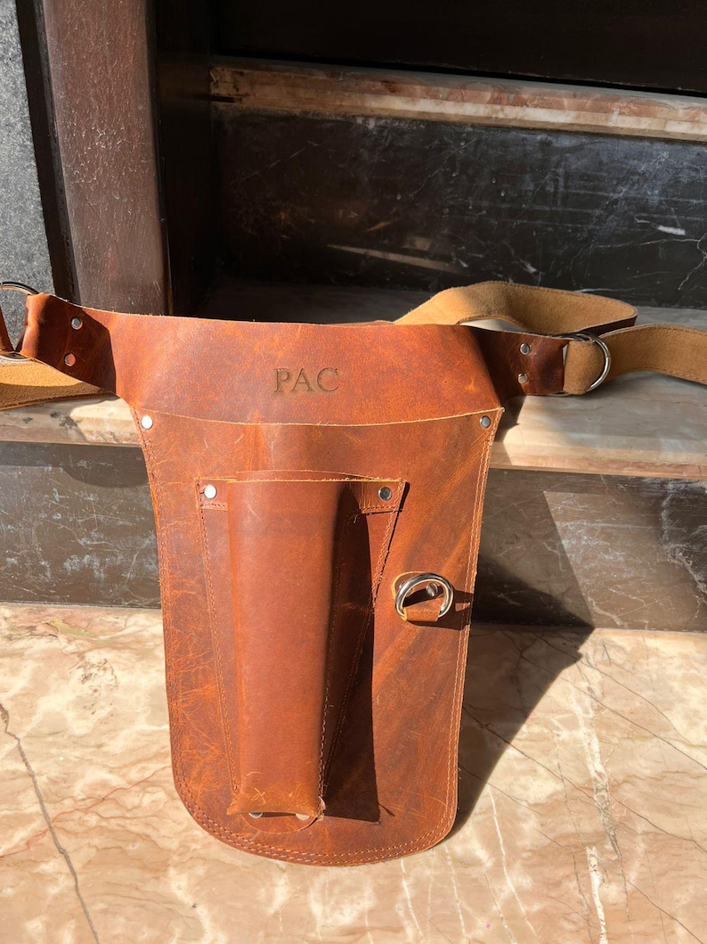 Puede incluir: Un cintur&oacute;n de herramientas de cuero marr&oacute;n con un solo bolsillo y un anillo de metal. El cintur&oacute;n tiene una correa que se coloca sobre el hombro y las palabras "PAC" est&aacute;n grabadas en la parte delantera.