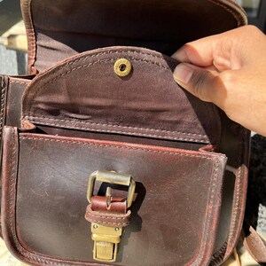 Peut inclure: Un sac &agrave; bandouli&egrave;re en cuir marron avec une boucle dor&eacute;e et un rabat avec un bouton dor&eacute;. Le sac est ouvert et l'int&eacute;rieur est doubl&eacute; de tissu marron.