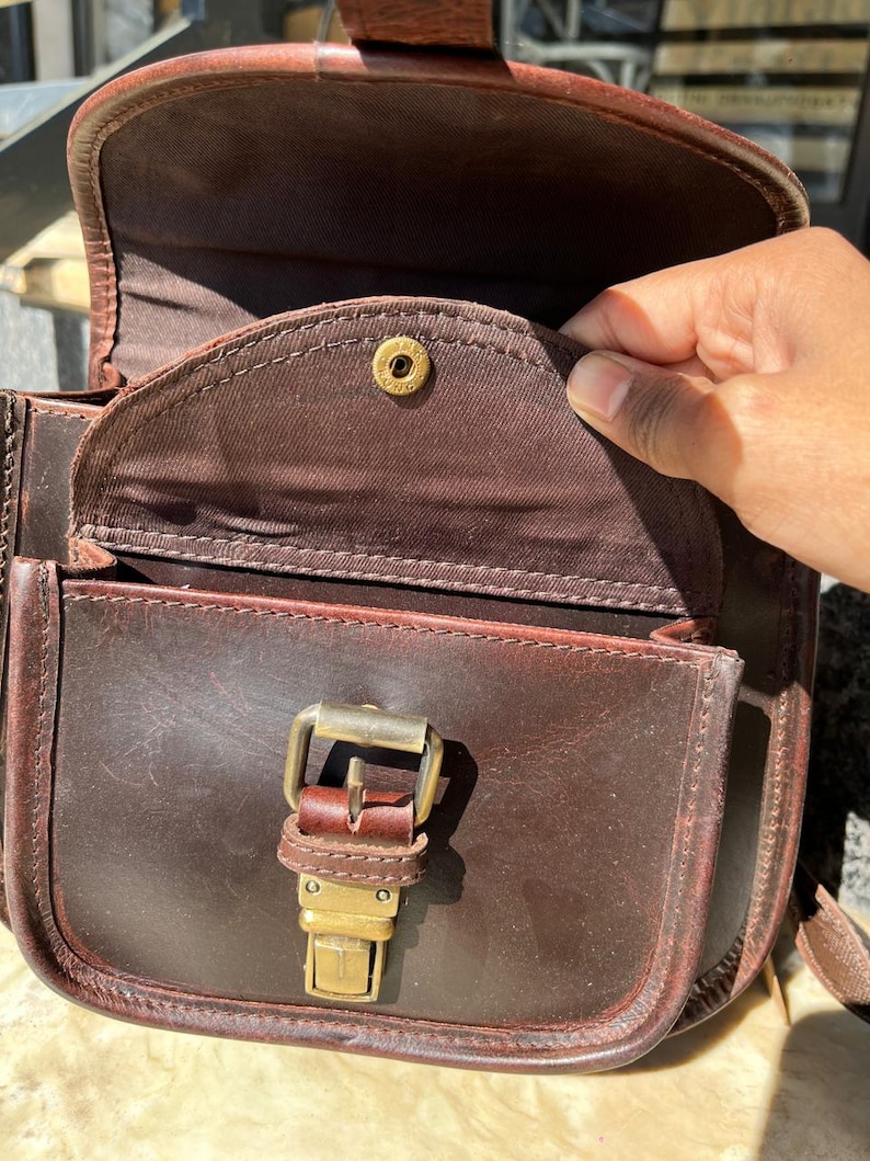 Peut inclure: Un sac &agrave; bandouli&egrave;re en cuir marron avec une boucle dor&eacute;e et un rabat avec un bouton dor&eacute;. Le sac est ouvert et l'int&eacute;rieur est doubl&eacute; de tissu marron.