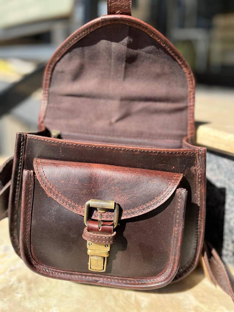 Peut inclure: Un sac &agrave; main en cuir marron avec une fermeture &agrave; rabat et une boucle en laiton. Le sac a une poche avant et une doublure en tissu marron.