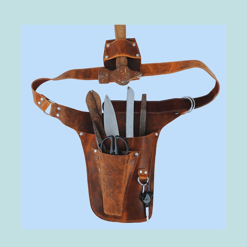 Puede incluir: Un cintur&oacute;n de herramientas de cuero marr&oacute;n con un martillo, tijeras y varias cuchillas. El cintur&oacute;n tiene un bolsillo grande en la parte delantera y un bolsillo m&aacute;s peque&ntilde;o en el lateral.