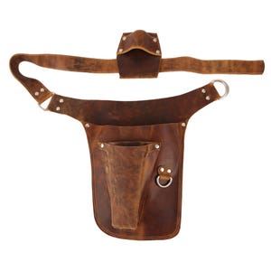 Puede incluir: Un cintur&oacute;n de herramientas de cuero marr&oacute;n con un bolsillo grande y un bolsillo m&aacute;s peque&ntilde;o en la parte delantera. El cintur&oacute;n tiene una hebilla de metal y dos lazos de metal.