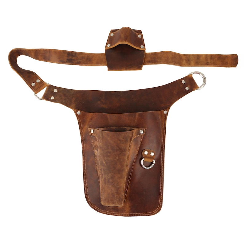 Puede incluir: Un cintur&oacute;n de herramientas de cuero marr&oacute;n con un bolsillo grande y un bolsillo m&aacute;s peque&ntilde;o en la parte delantera. El cintur&oacute;n tiene una hebilla de metal y dos lazos de metal.