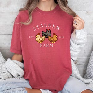 May include: A red t-shirt with a white graphic of a pixelated farm scene. The graphic says "Stardew Farm" with the text "Est. 2016" below the farm scene.