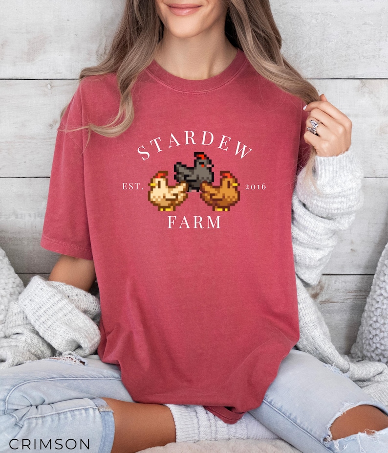 May include: A red t-shirt with a white graphic of a pixelated farm scene. The graphic says "Stardew Farm" with the text "Est. 2016" below the farm scene.
