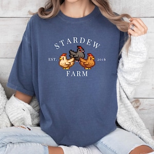 May include: A blue t-shirt with a white graphic of a farm scene. The graphic includes three chickens, two brown and one black, and the text "Stardew Farm Est. 2016".