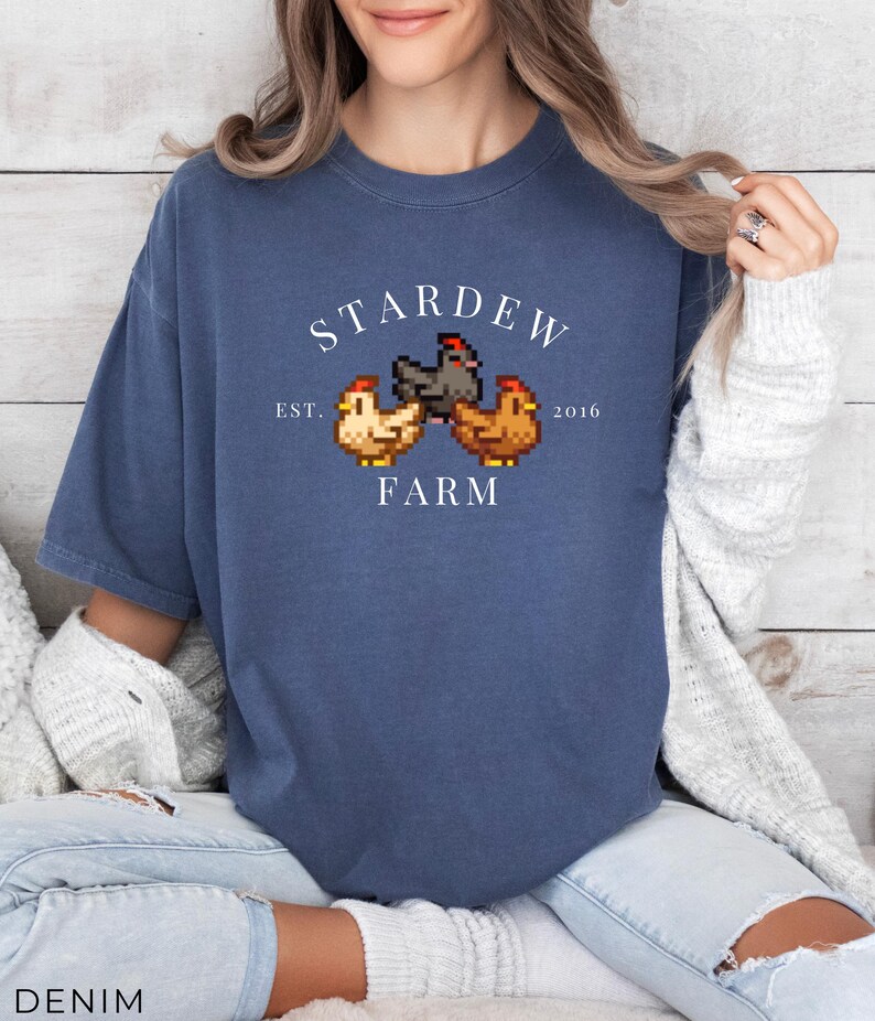 May include: A blue t-shirt with a white graphic of a farm scene. The graphic includes three chickens, two brown and one black, and the text "Stardew Farm Est. 2016".