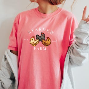 May include: A pink t-shirt with a white graphic of three pixelated chickens and the text "Stardew Farm Est. 2016".