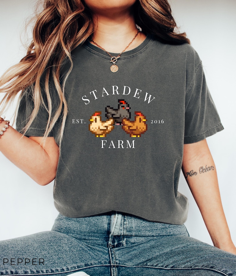 May include: A gray t-shirt with a white graphic of a video game logo. The logo says "Stardew Farm" with the text "Est. 2016" below it. The logo also features a pixelated image of three chickens.