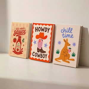May include: Three decorative books with colorful covers. The first book reads "this needs sauce" with a tomato sauce bottle illustration. The second book says "HOWDY COWBOY" with a cowboy boot and hat. The third book says "chill time" with a kangaroo illustration.
