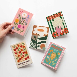 K&ouml;nnte beinhalten: Eine Sammlung von Gru&szlig;karten mit farbenfrohen Illustrationen. Eine Karte zeigt ein Blumendesign, eine andere eine Blume, und andere zeigen Pasta, Kirschen und Spaghetti. Die Karten liegen auf einer wei&szlig;en Oberfl&auml;che.