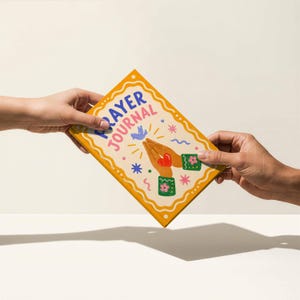 May include: A yellow and cream-colored prayer journal with the words "PRAYER JOURNAL" in blue and red. The cover features illustrations of praying hands, a dove, and floral accents. The journal is held by two hands.
