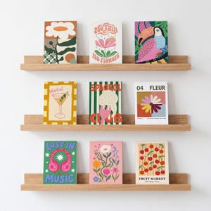 K&ouml;nnte beinhalten: Neun farbenfrohe Kunstdrucke auf Holzregalen vor einer wei&szlig;en Wand. Die Drucke zeigen verschiedene Illustrationen, darunter einen Martini, einen Papagei und die Worte "Lost in Music".