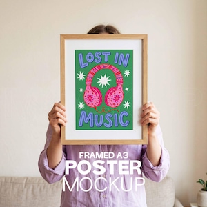 May include: A framed A3 poster mockup. The poster has a green background with the words "LOST IN MUSIC" in blue and pink headphones. The headphones have a decorative design. The poster is held up by a person.