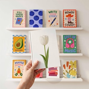 Pu&ograve; includere: Una collezione di diari colorati con vari disegni e testi, tra cui "Fill These Pages", "Write Your Story" e "Howdy Cowboy". I diari sono esposti su mensole bianche contro un muro bianco. Un diario presenta un tulipano bianco.