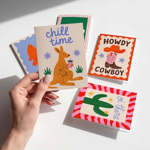 May include: A hand holds a card with a kangaroo illustration and the words "chill time." Other cards feature a fish, cactus, and cowboy boot designs. The cards have colorful illustrations and text on a white background.