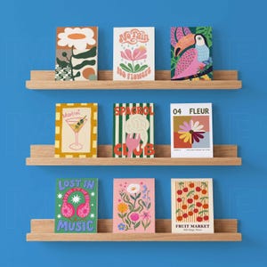 K&ouml;nnte beinhalten: Neun farbenfrohe Kunstdrucke, die auf Holzregalen vor einer blauen Wand ausgestellt sind. Die Drucke zeigen verschiedene Designs, darunter Blumenmuster, ein Martiniglas, einen Tukan und Texte wie "No Rain No Flowers" und "Lost in Music."